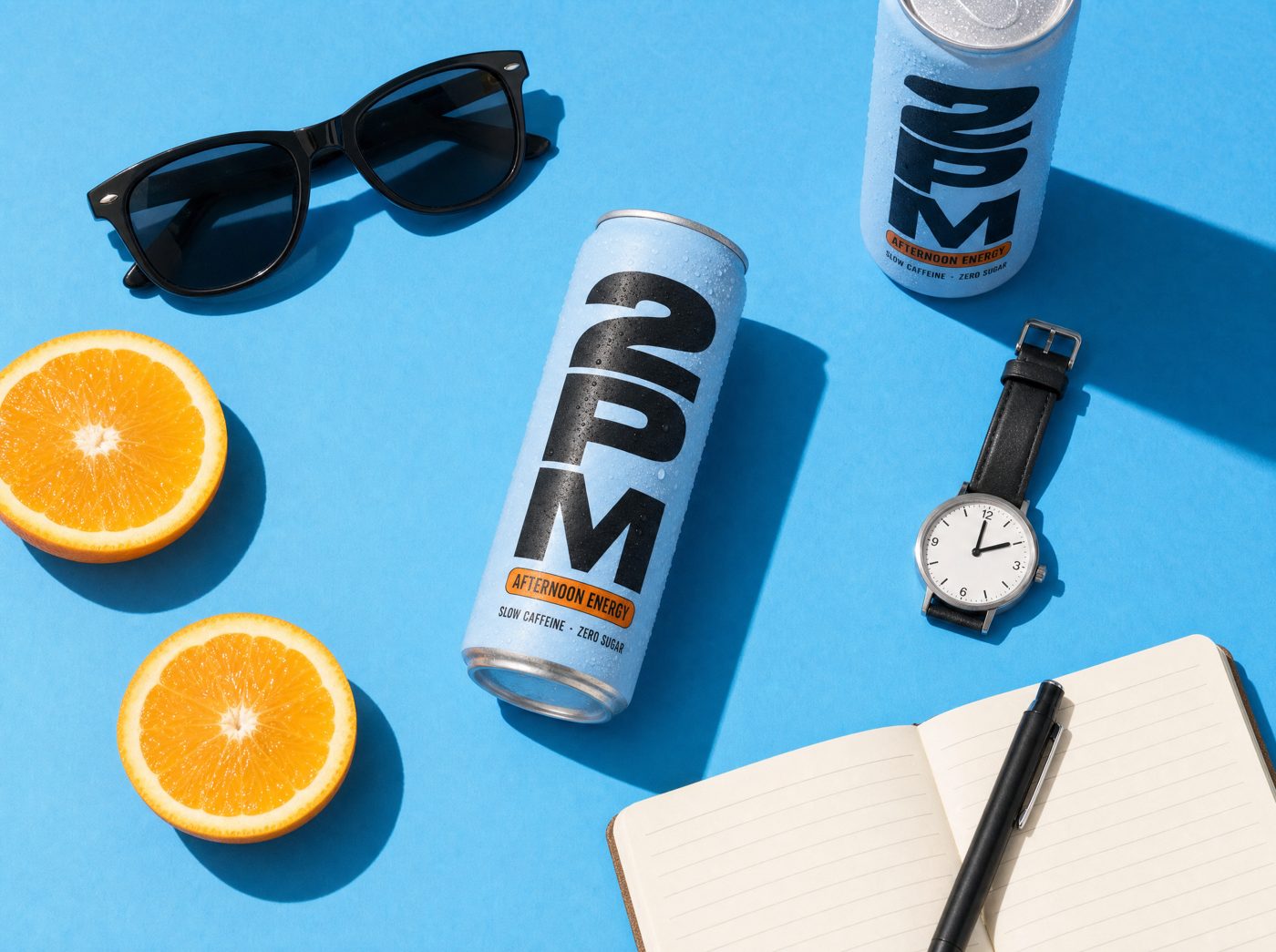 A can of 2PM on a bright blue surface with orange slices, sunglasses and a watch reading 2:00